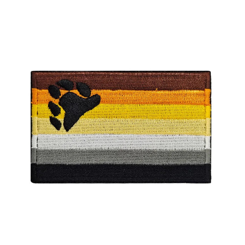 Gestickter Aufnäher mit der Bären-Flagge, die braune, gelbe, orange, weiße, graue und schwarze Streifen zeigt, mit einem schwarzen Bärentatzen-Motiv in der oberen linken Ecke.