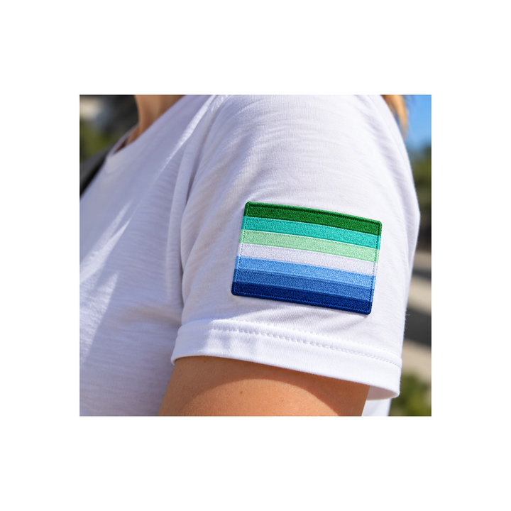 Gestickter Gay Men Pride Patch mit sieben Streifen in Grün, Türkis, Weiss und Blau auf dem Oberarm eines weissen T-Shirts. Pride-Aufnäher, 8 × 5 cm.