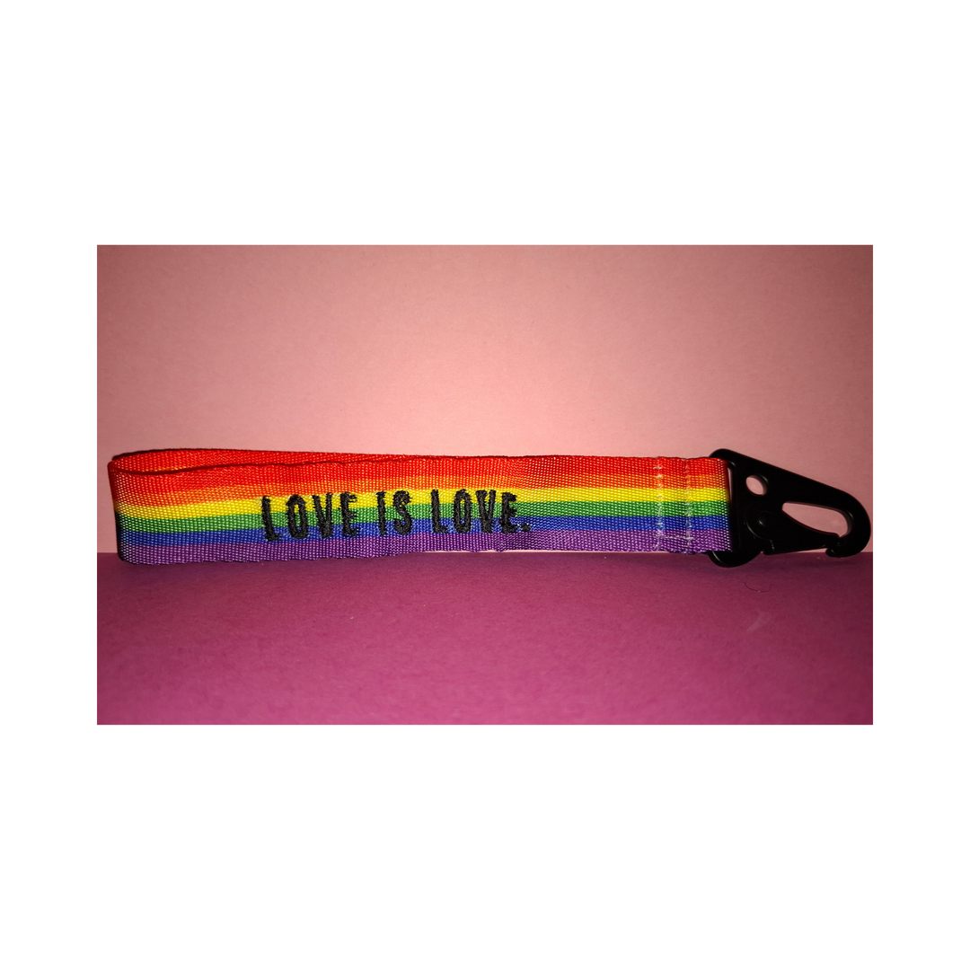 Regenbogen-Schlüsselanhänger mit schwarzem Karabiner und der Aufschrift „LOVE IS LOVE“. Farbenfrohes LGBTQIA+ Pride Accessoire aus robustem Gurtband auf rosafarbenem Hintergrund.