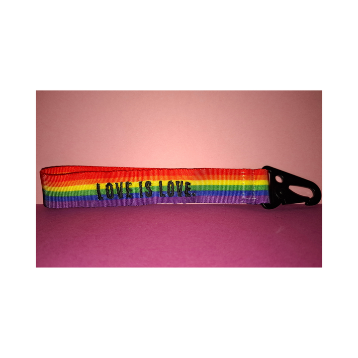 Regenbogen-Schlüsselanhänger mit schwarzem Karabiner und der Aufschrift „LOVE IS LOVE“. Farbenfrohes LGBTQIA+ Pride Accessoire aus robustem Gurtband auf rosafarbenem Hintergrund.