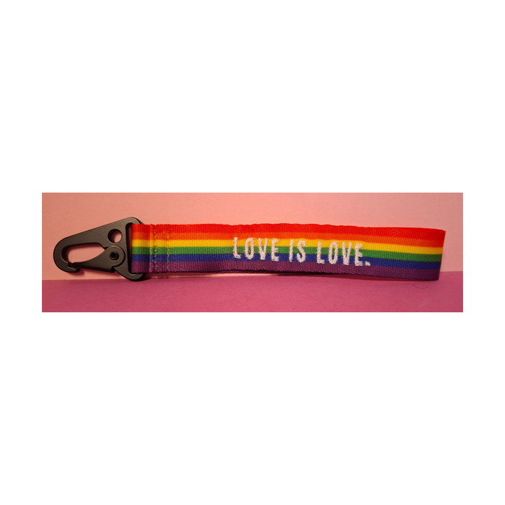 Regenbogen-Schlüsselanhänger mit schwarzem Karabiner und der Aufschrift „LOVE IS LOVE“. Farbenfrohes LGBTQIA+ Pride Accessoire aus robustem Gurtband auf rosafarbenem Hintergrund.