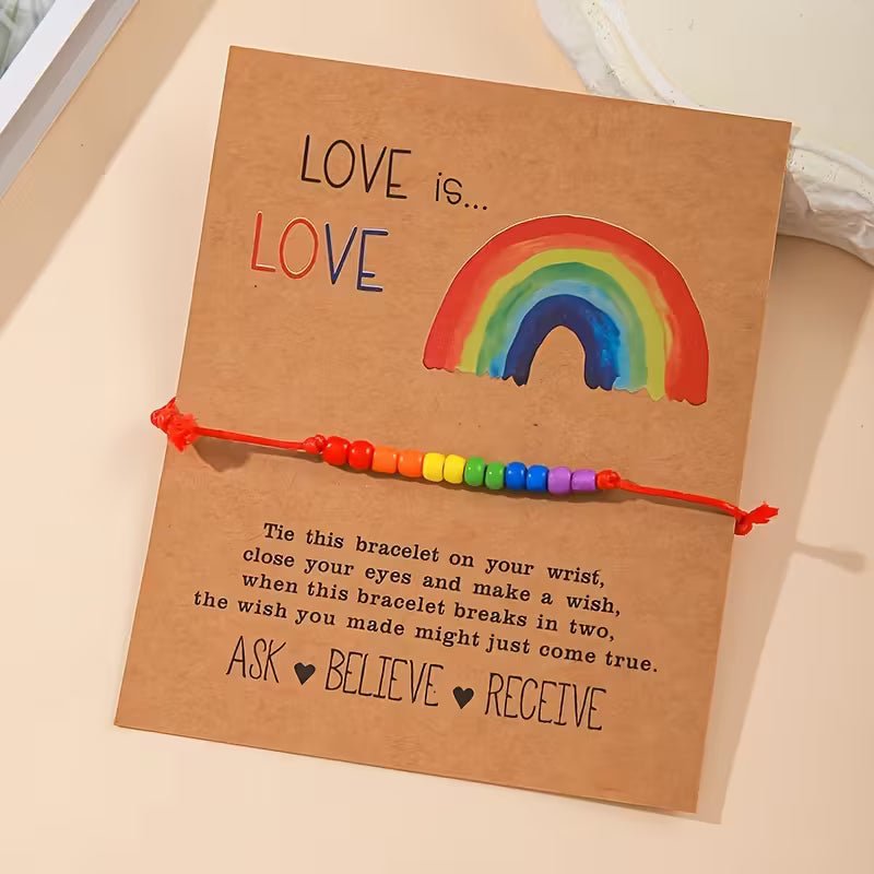 Love-is-Love Armband mit roter Schnur und Regenbogen-Perlen, befestigt auf brauner Kartonkarte mit Regenbogen-Illustration