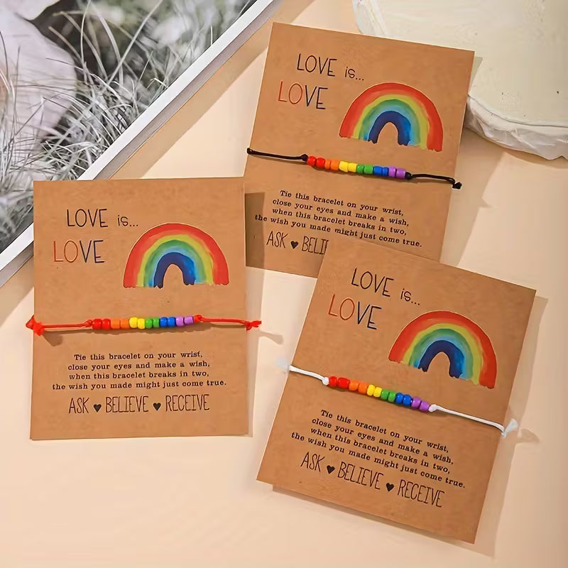 Drei Love-is-Love Armbänder mit Regenbogenperlen auf braunen Karten, jeweils mit roter, schwarzer und weißer Schnur