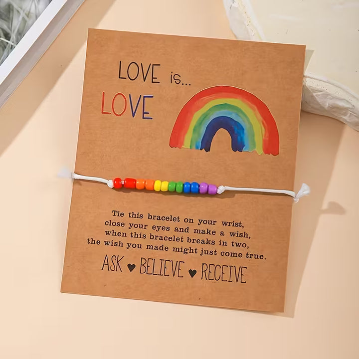 Love-is-Love Armband mit weißer Schnur und Regenbogen-Perlen, befestigt auf brauner Kartonkarte mit Regenbogenillustration