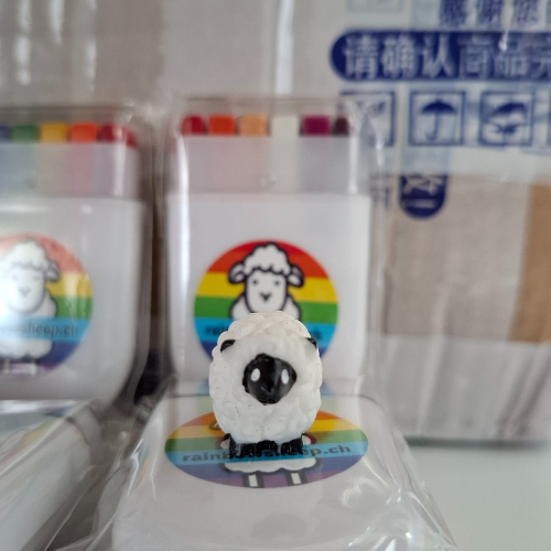 Mehrere weisse Etuis mit Schminkstiften in Regenbogen- und Lesben-Flaggenfarben. Auf jedem Etui ist ein runder Aufkleber mit einem Schaf vor Regenbogenhintergrund und der Aufschrift rainbowsheep.ch. Im Vordergrund steht eine kleine Schaf-Figur aus Kunststoff. Im Hintergrund ist ein Karton mit blauen Aufdrucken zu sehen.