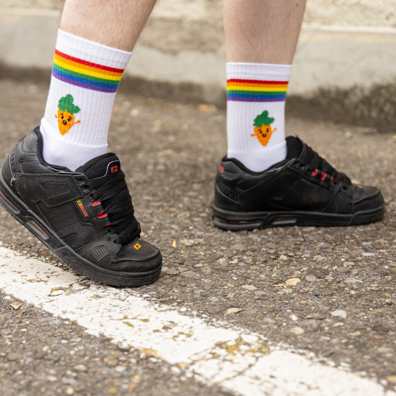 Person trägt offizielle Pride Aargau Rüebli Socken mit Regenbogenstreifen und Rüebli Motiv in schwarzen Schuhen