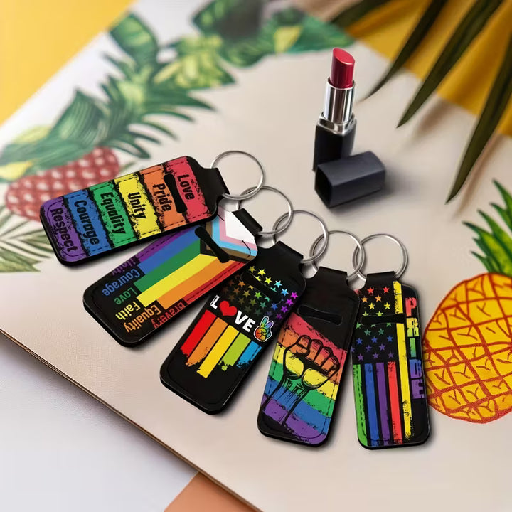Pride-Lippenpflegestift-Halter im Regenbogen-Design mit Schlüsselringen, neben Lippenstift und Deko platziert.