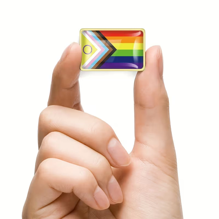Kleiner rechteckiger Progress Pride Flag Pin mit goldfarbenem Rand, zwischen zwei Fingern gehalten, Stickeroberfläche, nicht wasserdicht