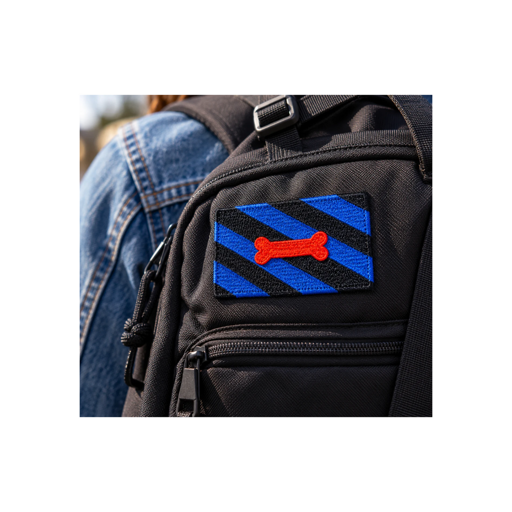 Blau-schwarzer Puppy Play Patch mit rotem Knochenmotiv auf einem schwarzen Rucksack. Gestickter Aufnäher, 8 × 5 cm, ohne Klettverschluss.