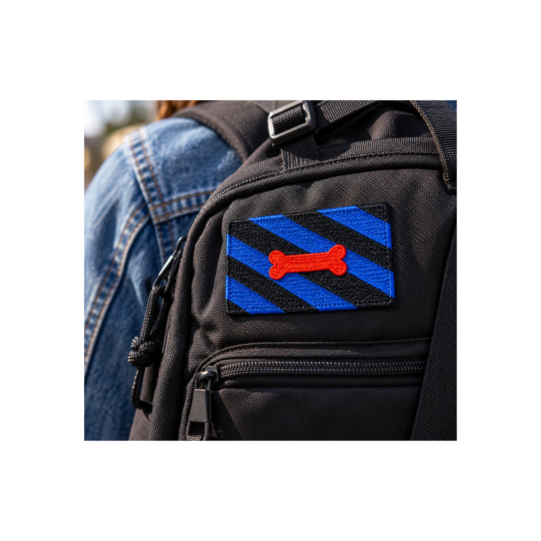 Blau-schwarzer Puppy Play Patch mit rotem Knochenmotiv auf einem schwarzen Rucksack. Gestickter Aufnäher, 8 × 5 cm, ohne Klettverschluss.