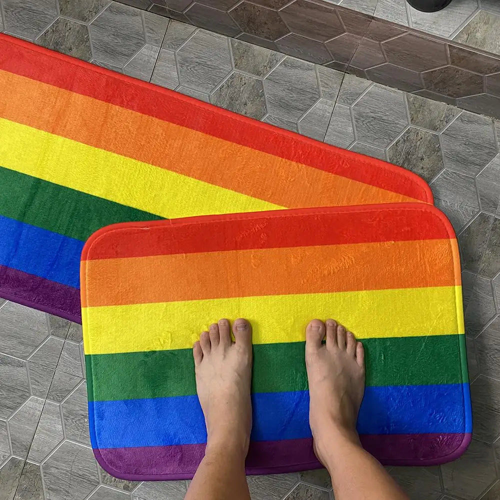 Zwei Fußmatten mit Regenbogenflaggen-Design, auf einer stehenden nackten Füße, platziert auf Fliesenboden.