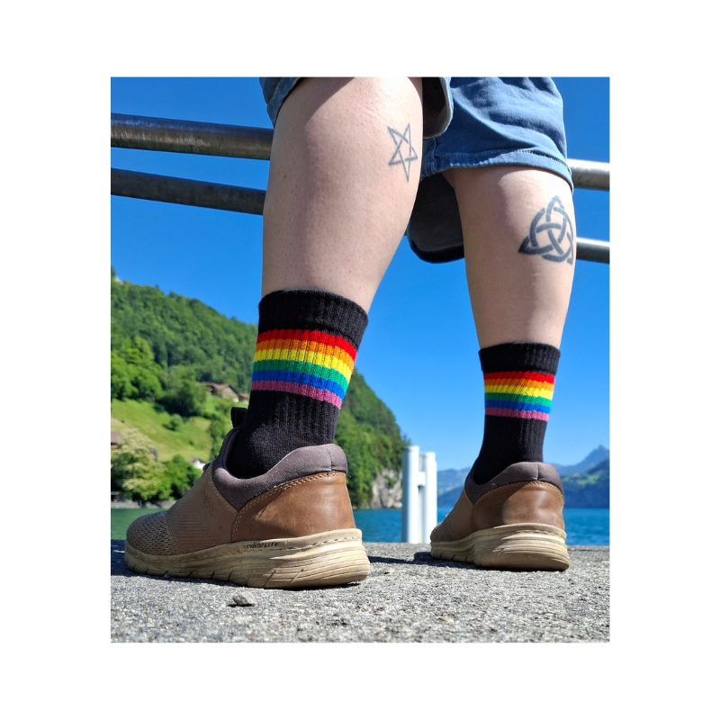 Person trägt schwarze Regenbogen Pride Socken mit bunten Streifen am Seeufer bei sonnigem Wetter