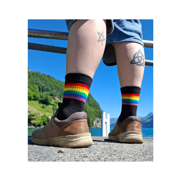 Person trägt schwarze Regenbogen Pride Socken mit bunten Streifen am Seeufer bei sonnigem Wetter