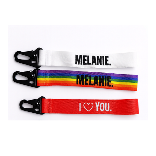 Vier bunte Schlüsselanhänger aus Stoff mit Karabinerverschluss. Der obere ist weiß mit dem Schriftzug "MELANIE.", der zweite hat ein Regenbogenmuster mit ebenfalls "MELANIE.", der dritte ist rot mit "I ❤️ YOU.", und der untere ist blau mit einem Symbol für weibliche Gleichberechtigung.