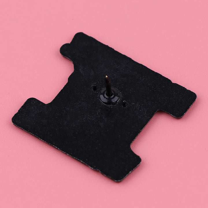 Rückseite eines schwarzen Emaille-Pins mit Metallnadel auf rosa Hintergrund