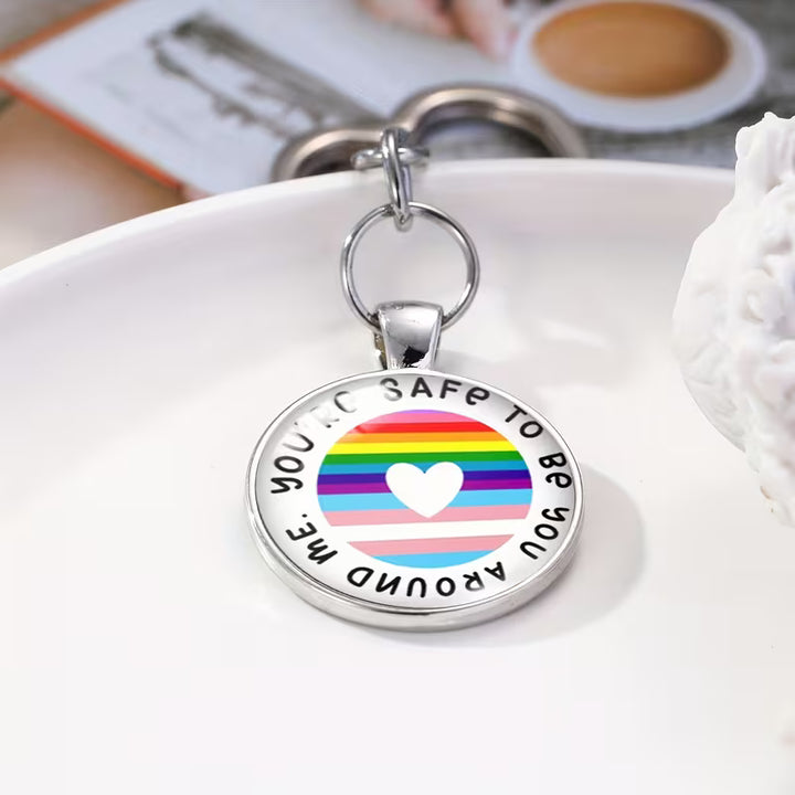 Schlüsselanhänger mit herzförmigem Metallring und rundem Anhänger mit Regenbogenstreifen und weißem Herz in der Mitte; Text auf dem Anhänger: ‘You’re safe to be around me’.