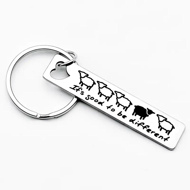 Metall-Schlüsselanhänger „It’s good to be different“ mit fünf Schafen, eines schwarz, auf weißem Hintergrund