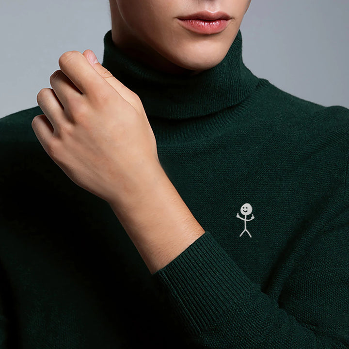 Person in grünem Rollkragenpullover mit silbernem Strichmännchen-Pin auf der Brust vor neutralem Hintergrund.