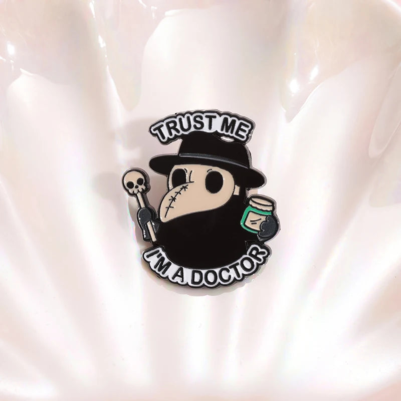 Ein lustiger Ansteck-Pin mit einem niedlichen Pestdoktor in typischer schwarzer Kleidung und Schnabelmaske. Er hält einen kleinen Totenkopf-Stab und ein Medizinfläschchen, während der Text „Trust Me, I'm a Doctor“ darüber und darunter steht. Perfekt für Fans von Gothic-Ästhetik, dunklem Humor oder historischen Medizin-Motiven.